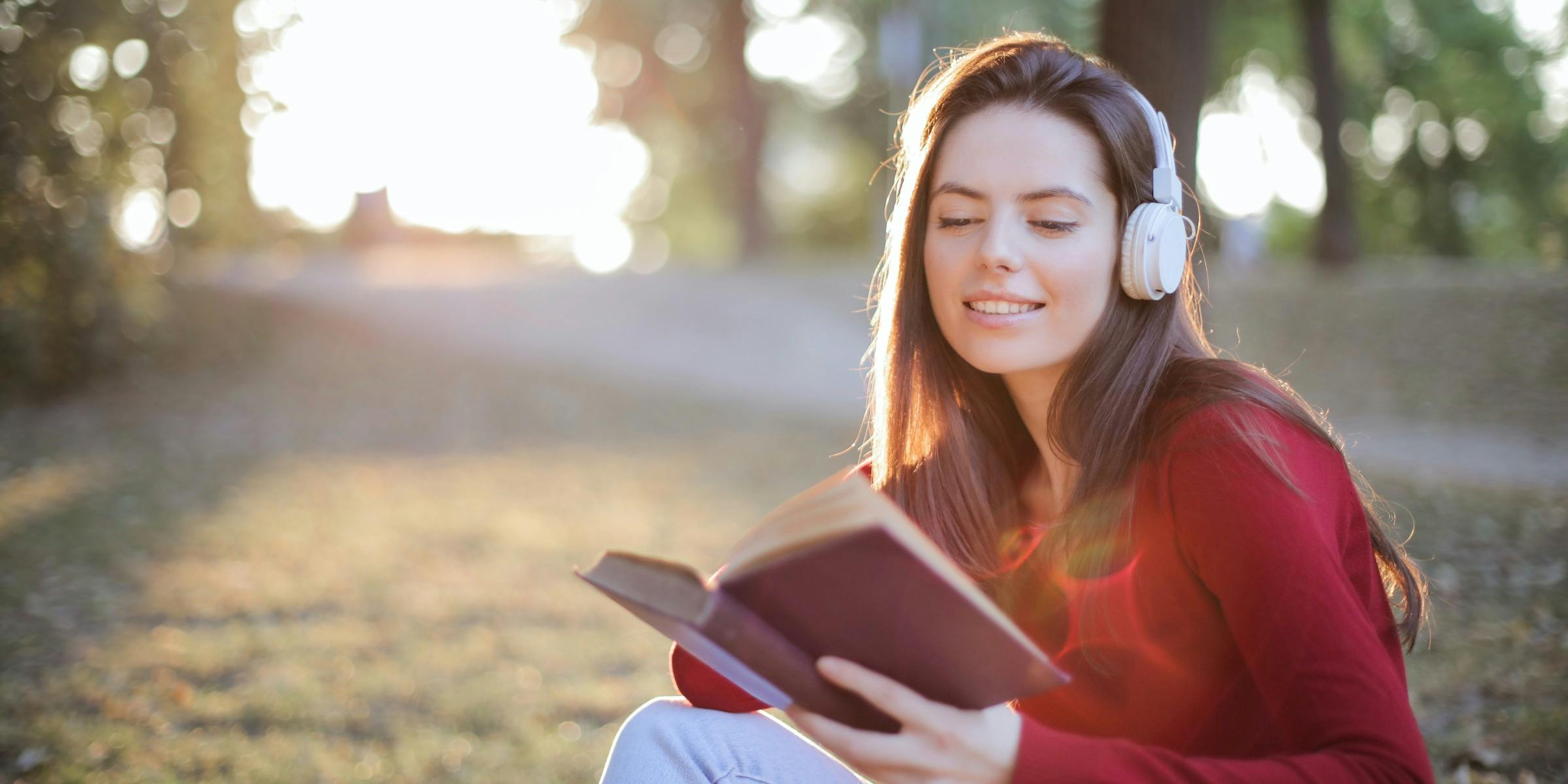 Young woman in a field wearing headphones while reading a book.